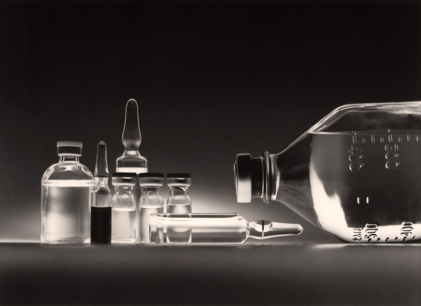 Black-and-white still life of laboratory ampoules, vials and glassware arranged on a dark surface.