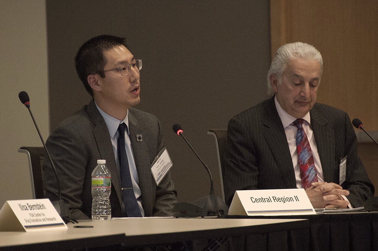 Two speakers at a microphone-equipped table with name placards during an FDA pharmacy compounding advisory meeting.