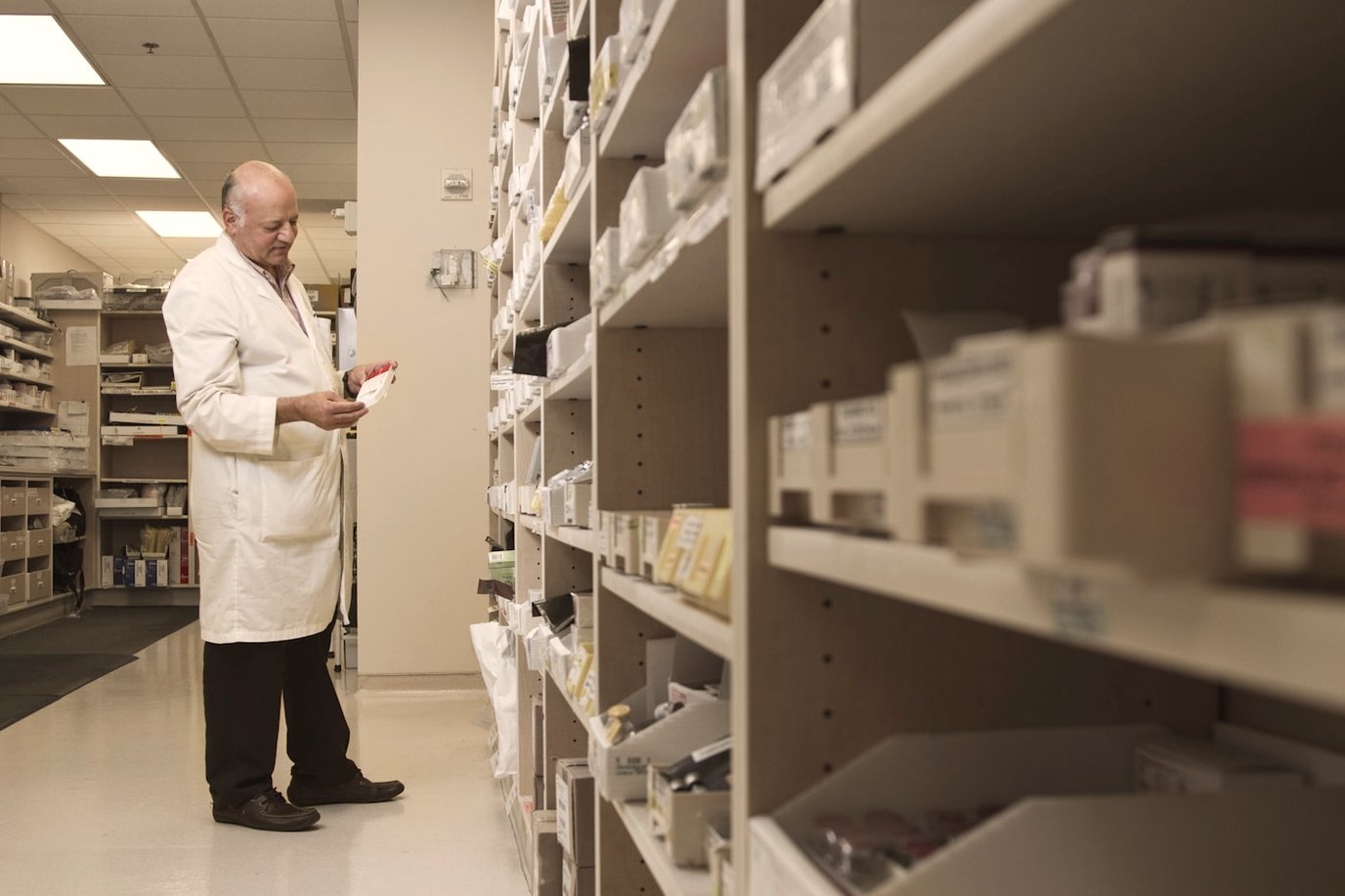 Pharmacist in a white coat working at pharmacy shelving stocked with medication packages.