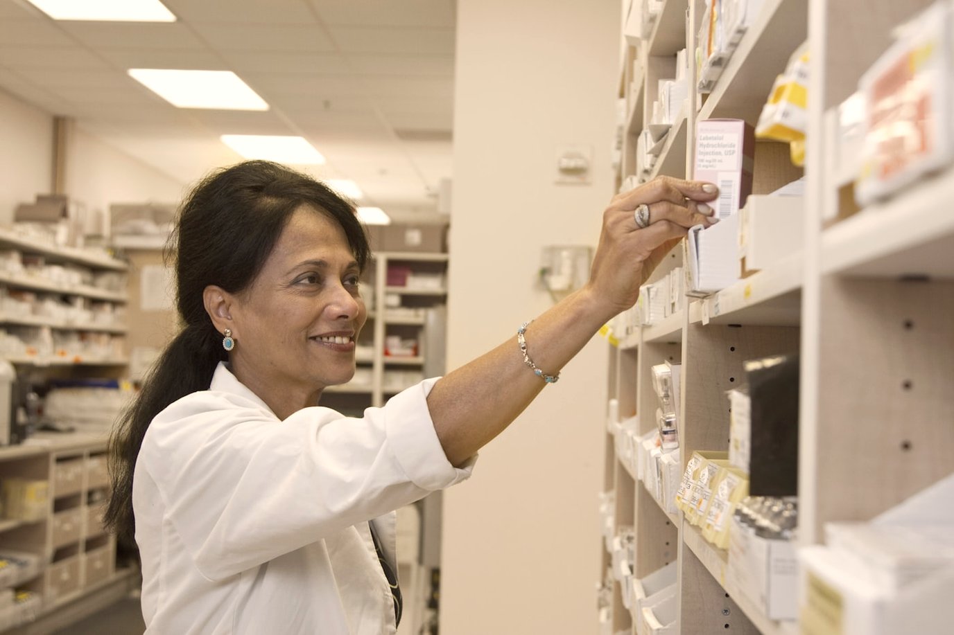 Pharmacist reaching for stock on a wall of pharmacy shelving filled with labelled boxes.