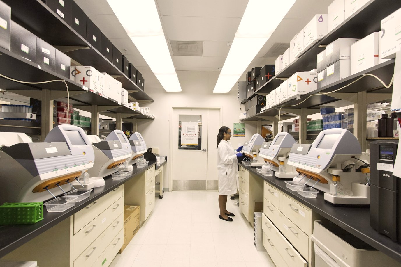 Long research laboratory bench lined with analytical instruments, a technician in a lab coat working at the far end.